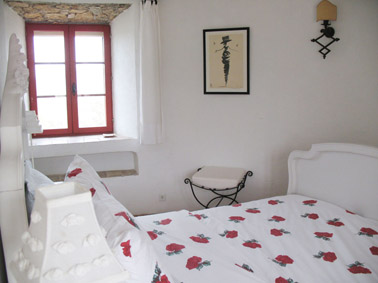 The bedroom on the first floor of Moulin Tower showing antique white wooden bed with white table lamps, wrought iron chair and wrought iron wall light. There is a window with a view of the valley.